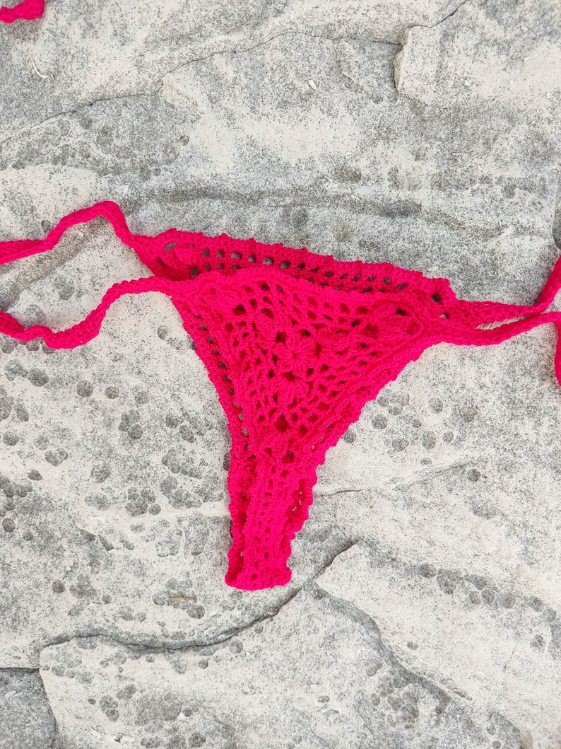 May include: A bright pink, handmade crochet bikini bottom. The bottom features a delicate, open weave design and string ties. The garment is displayed on a textured, gray stone surface. The crochet work appears to be of a medium weight yarn.