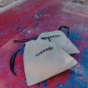 May include: Two white drawstring bags with black drawstrings and the text "creaspir" printed in black on each bag. The bags are on a red and blue surface.