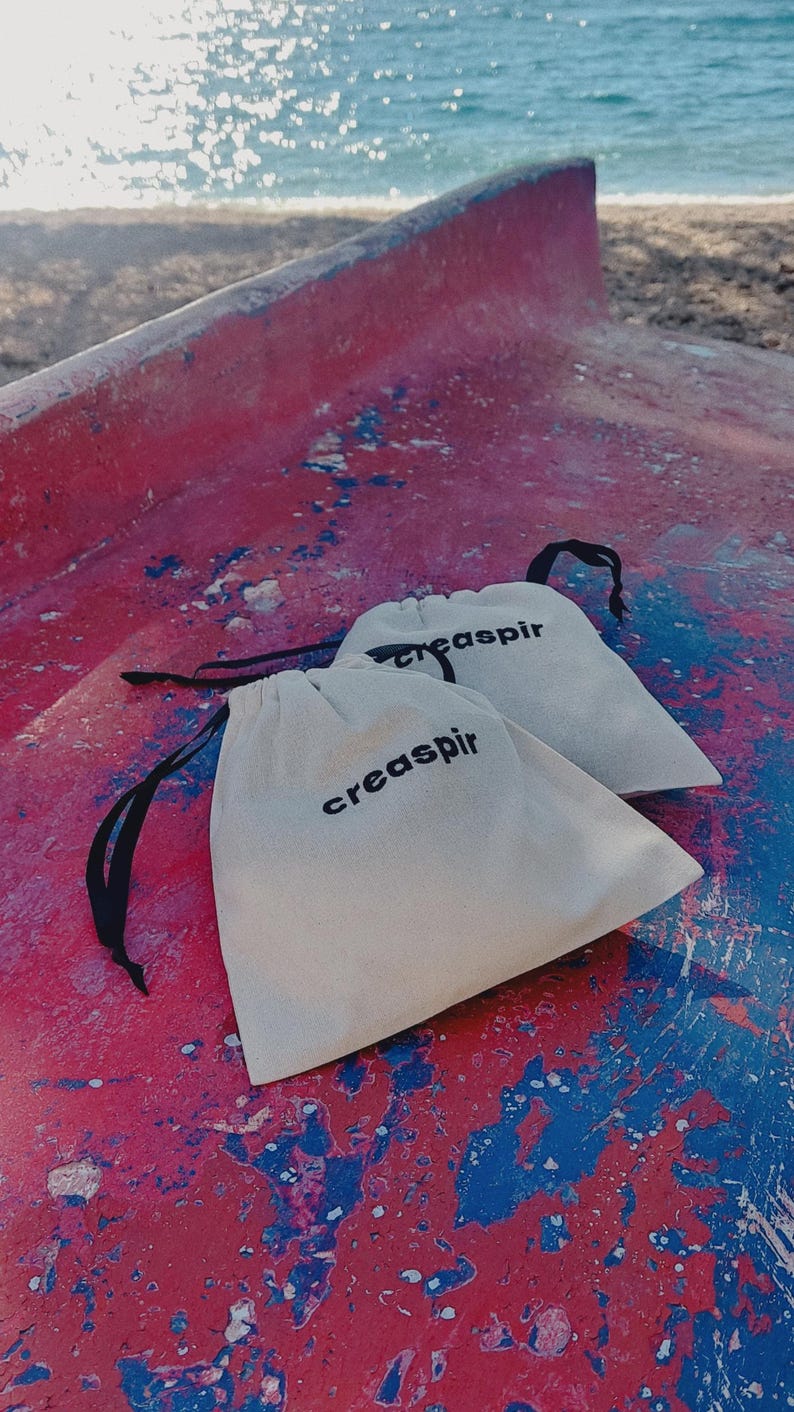 May include: Two white drawstring bags with black drawstrings and the text "creaspir" printed in black on each bag. The bags are on a red and blue surface.