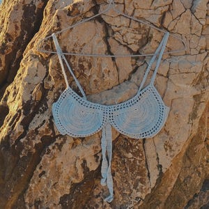May include: A light blue crocheted bikini top hanging on a wire hanger against a rocky background.