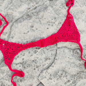 May include: A bright pink, handmade crochet bikini top. The top features a triangle design with a halter neck and back ties. The open weave of the crochet creates a delicate, airy look. The bikini top is displayed on a textured, gray stone surface.
