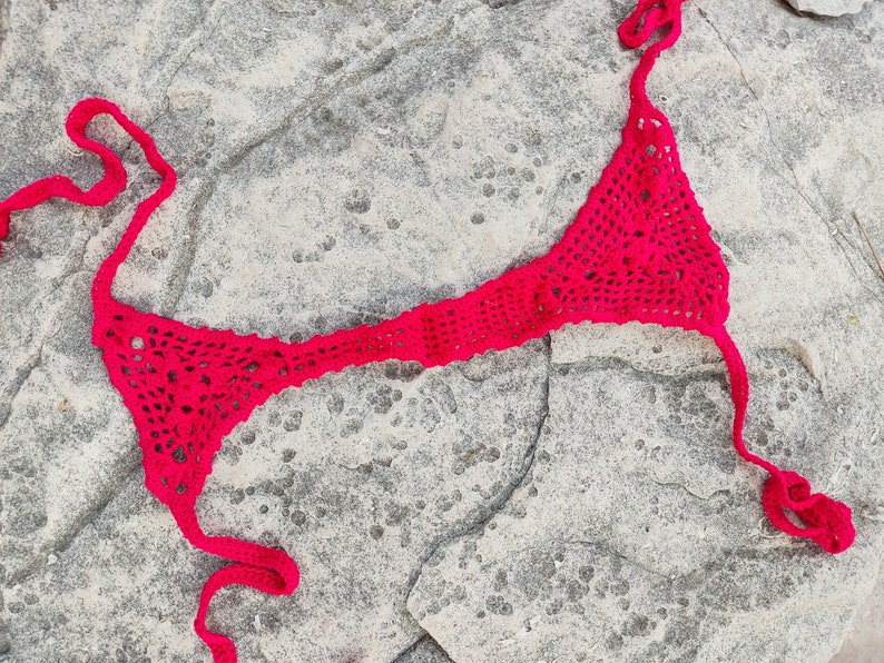 May include: A bright pink, handmade crochet bikini top. The top features a triangle design with a halter neck and back ties. The open weave of the crochet creates a delicate, airy look. The bikini top is displayed on a textured, gray stone surface.