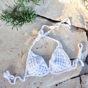 May include: White crochet bikini top with a scalloped design. The top is lying on a light gray rocky surface.