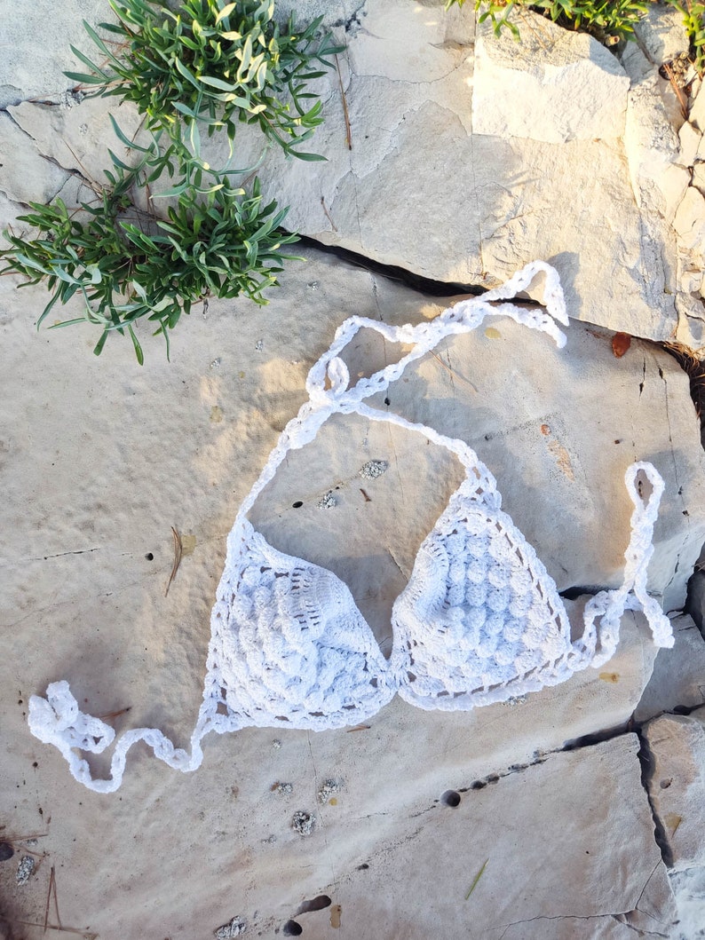 May include: White crochet bikini top with a scalloped design. The top is lying on a light gray rocky surface.