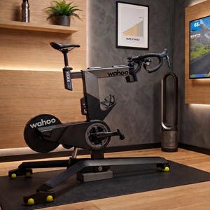 May include: A Wahoo KICKR smart indoor cycling trainer featuring a black and grey frame, drop handlebars, a saddle, and a flywheel with the Wahoo logo. The unit is positioned on a black mat with yellow caps on the base feet, designed for stationary cycling.
