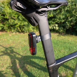 May include: A black bicycle seat post featuring a mounted red rear safety light and a small black zippered saddle bag attached directly underneath the bicycle seat. The light is secured to the post with a black rubber strap and metal hardware.