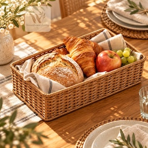 May include: A rectangular woven basket with a light brown finish, lined with a beige fabric featuring a thin dark stripe. The basket contains a round loaf of bread, two golden-brown croissants, a red apple, and a cluster of green grapes.