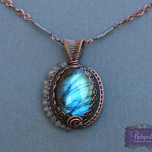 以下が含まれることがあります： 銅線で巻かれたペンダントで、大きな楕円形の青いラブラドライトの宝石が特徴です。ペンダントは、より小さな透明なファセットカットの石でアクセントが付けられており、銅のチェーンからぶら下がっています。