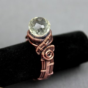 Pode incluir: Um anel envolto em fio de cobre com uma gema facetada transparente. O anel está em um suporte de veludo preto.