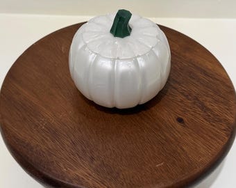 Puede incluir: Un recipiente con tapa en forma de calabaza de color blanco nacarado, con un tallo de color verde oscuro como asa. El objeto presenta estrías verticales y un acabado liso y brillante, con una forma redondeada y segmentada de calabaza.