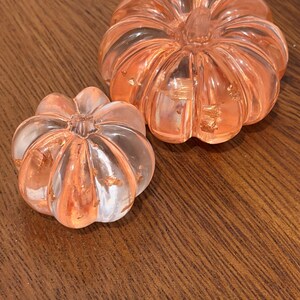 May include: Two clear, ribbed pumpkin-shaped ornaments made of resin, each featuring embedded metallic copper-colored flakes throughout the material. The larger pumpkin is positioned behind the smaller one, both displaying a glossy finish.