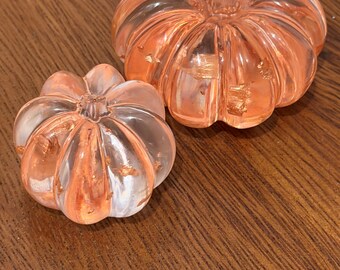Puede incluir: Dos adornos transparentes con forma de calabaza acanalada hechos de resina, cada uno con escamas metálicas de color cobre incrustadas en el material. La calabaza más grande está colocada detrás de la más pequeña.