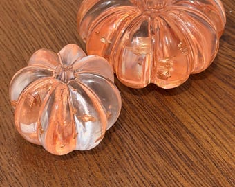 Puede incluir: Dos adornos transparentes con forma de calabaza acanalada hechos de resina, cada uno con escamas metálicas de color cobre incrustadas en el material. La calabaza más grande está colocada detrás de la más pequeña.