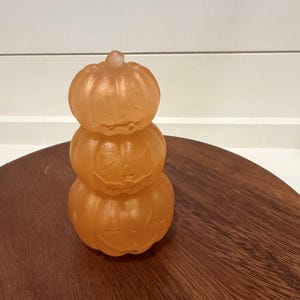 May include: A stack of three orange, translucent, pumpkin-shaped objects. Each pumpkin features a carved jack-o'-lantern face with triangular eyes, a nose, and a jagged mouth. The surface has a smooth, slightly frosted texture throughout.