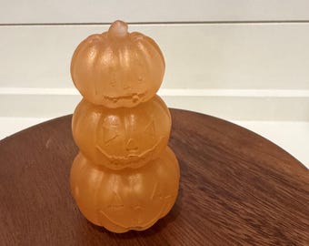 Puede incluir: Una pila de tres objetos de color naranja, translúcidos y con forma de calabaza. Cada calabaza presenta una cara de linterna tallada con ojos triangulares, una nariz y una boca dentada. La superficie tiene una textura lisa y ligeramente esmerilada.