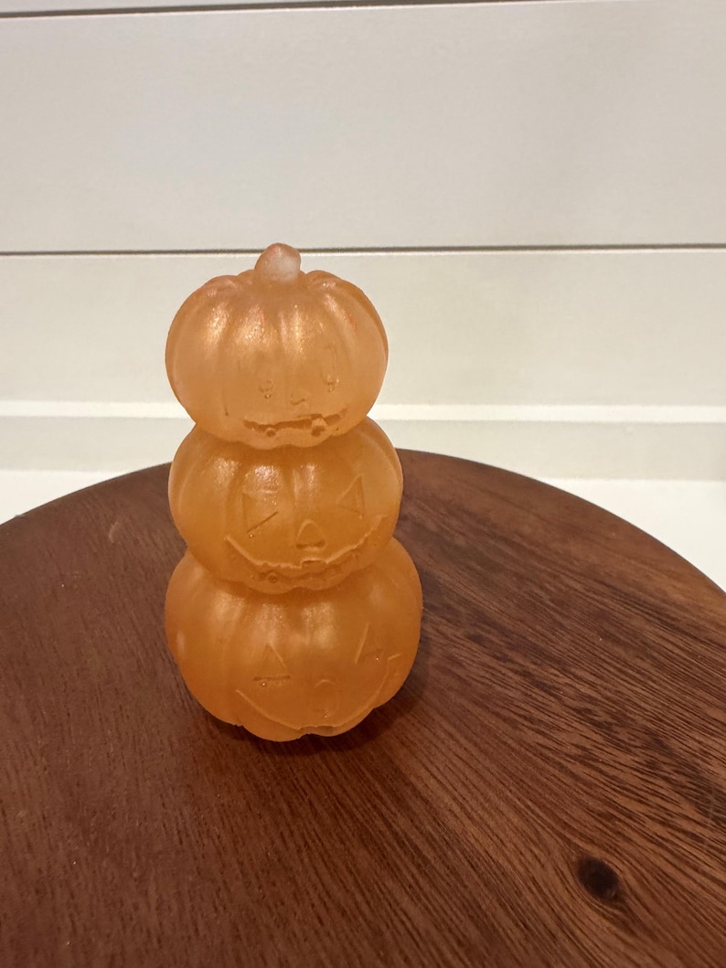 Puede incluir: Una pila de tres objetos de color naranja, transl&uacute;cidos y con forma de calabaza. Cada calabaza presenta una cara de linterna tallada con ojos triangulares, una nariz y una boca dentada. La superficie tiene una textura lisa y ligeramente esmerilada.