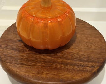 Puede incluir: Un recipiente translúcido de color naranja con forma de calabaza, con tapa a juego y un tallo central, que presenta un acabado liso y brillante con crestas verticales visibles, apoyado sobre un soporte circular de madera con tres patas cortas.