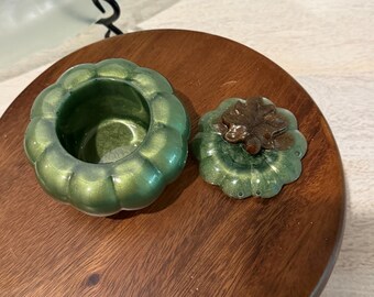 Puede incluir: Un pequeño recipiente redondo con forma de calabaza, acabado en verde metálico y tapa separada. La tapa presenta un asa marrón esculpida en forma de hoja. El recipiente está abierto, mostrando una superficie interior verde a juego.