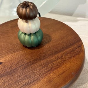 May include: A stack of three small, smooth, rounded pumpkin-shaped ornaments. The top pumpkin is metallic bronze, the middle is pearlescent white, and the bottom is metallic green. They are resting on a circular, polished dark wood surface.