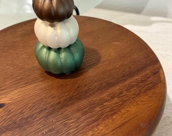 Puede incluir: Una pila de tres pequeños adornos lisos con forma de calabaza. La calabaza superior es de color bronce metálico, la del medio es de color blanco nacarado y la inferior es de color verde metálico. Descansan sobre una superficie circular de madera oscura.