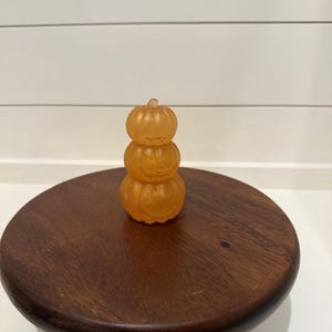 Puede incluir: Una pila de tres calabazas naranjas transl&uacute;cidas hechas de un material liso. Cada calabaza presenta una cara de linterna tallada con ojos y boca simples. Las calabazas est&aacute;n dispuestas verticalmente, con la m&aacute;s peque&ntilde;a arriba y la m&aacute;s grande abajo, creando una sola figura decorativa.