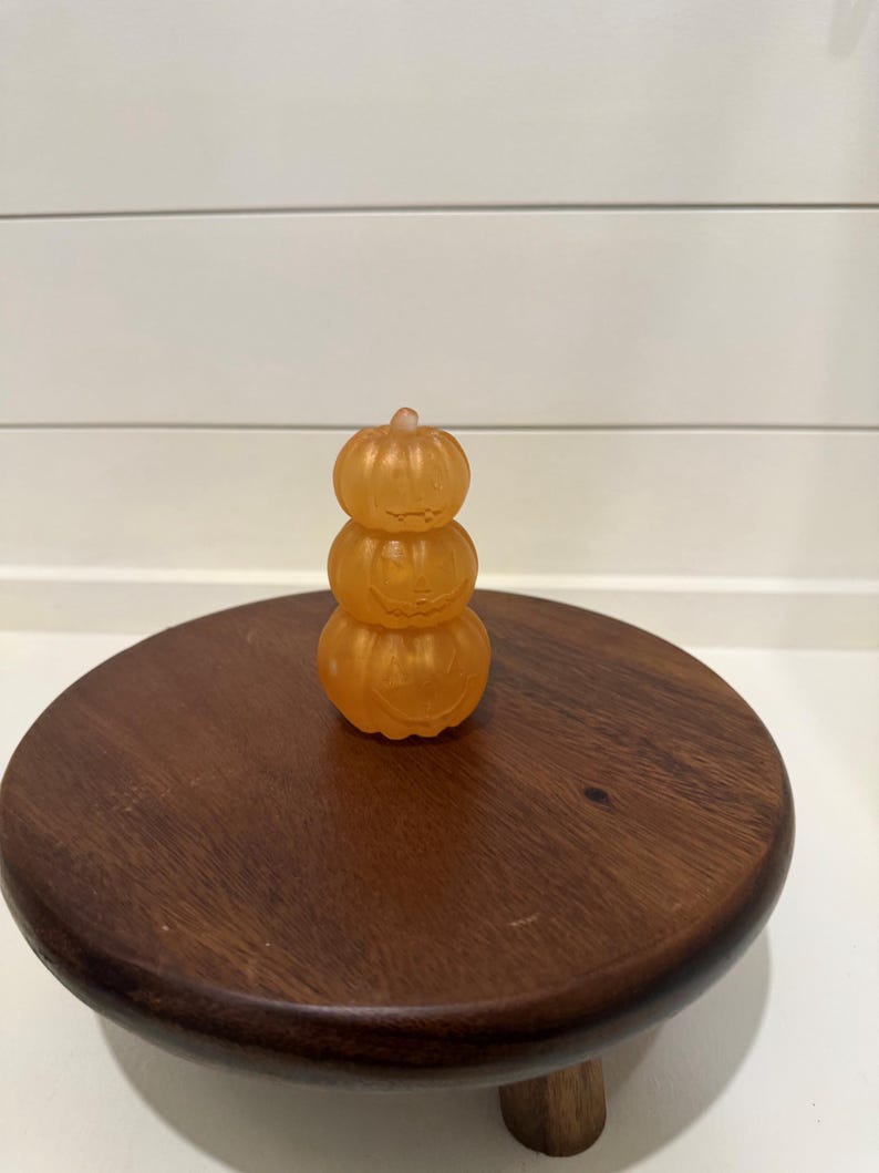 Puede incluir: Una pila de tres calabazas naranjas transl&uacute;cidas hechas de un material liso. Cada calabaza presenta una cara de linterna tallada con ojos y boca simples. Las calabazas est&aacute;n dispuestas verticalmente, con la m&aacute;s peque&ntilde;a arriba y la m&aacute;s grande abajo, creando una sola figura decorativa.
