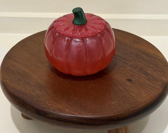 Puede incluir: Un recipiente de vidrio rojo translúcido con forma de calabaza y tapa, que presenta un tallo verde oscuro como asa. La calabaza tiene crestas verticales y un acabado liso, apoyada sobre un soporte de madera circular con tres patas.