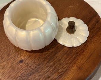 Puede incluir: Un recipiente blanco nacarado con forma de calabaza y su tapa a juego. La tapa presenta un asa oscura en forma de tallo. El recipiente tiene un exterior acanalado y un acabado liso y brillante, presentado como un conjunto de dos piezas.