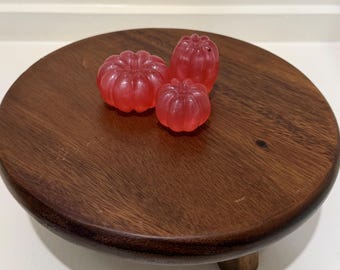 Puede incluir: Tres objetos pequeños, translúcidos y de color rosa con forma de calabaza y textura acanalada, dispuestos sobre un soporte redondo de madera marrón oscuro con patas cortas. Los objetos tienen un acabado liso y brillante.