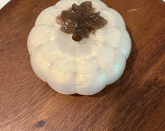 Puede incluir: Un objeto con forma de calabaza de color blanco nacarado con una superficie lisa y redondeada y un sutil brillo dorado. La parte superior presenta un detalle de tallo en forma de hoja de color marrón oscuro y brillante.