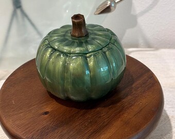 Puede incluir: Un pequeño recipiente de cerámica en forma de calabaza de color verde con un acabado metálico brillante y un tallo marrón oscuro. La calabaza presenta crestas verticales y una tapa extraíble, con una superficie lisa y reflectante.