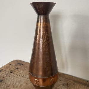 Könnte beinhalten: Eine hohe, konische Metallvase mit dunkelbraunem, gebürstetem Finish. Der Körper weist ein horizontales Band nahe der Basis mit kreisförmigen Prägedetails auf. Der Hals weitet sich nach oben hin aus und die Oberfläche hat eine texturierte Optik.