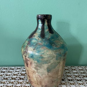 以下が含まれることがあります： 丸みを帯びた胴体と細い首を持つ陶器製の花瓶。ターコイズ、クリーム、ブラウンの色合いのまだら模様の釉薬が施されています。上部の縁はダークブラウンの垂れ釉で仕上げられており、質感のある表面に流れ落ちるようなデザインです。