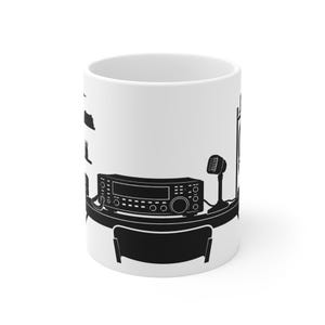 May include: A white ceramic mug featuring a black graphic illustration of a radio station setup. The design includes a desk with a radio transceiver, a microphone, and a chair, with additional radio equipment silhouettes on the side.