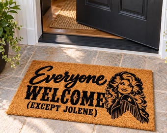 May include: A rectangular tan coir doormat featuring black text that reads Everyone WELCOME (EXCEPT JOLENE) alongside a black graphic illustration of a woman with voluminous hair wearing a star-patterned shirt with fringe detailing.