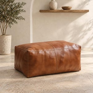 May include: A rectangular leather ottoman in a warm tan brown colour, featuring visible light-coloured stitching along the seams and a smooth, slightly textured surface. The piece has a structured, low-profile design with rounded edges.