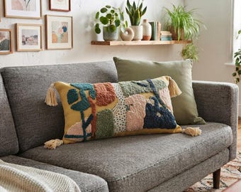 Può includere: Un cuscino rettangolare con base gialla e forme astratte trapuntate multicolori in verde ottanio, verde salvia, terracotta e crema. Il cuscino è arricchito da nappe color crema su ciascuno dei quattro angoli per una maggiore consistenza.