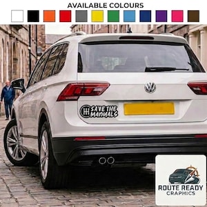 May include: A white vinyl decal applied to the rear of a white SUV, featuring a black gear shift pattern icon and the text "SAVE THE MANUALS" in a bold, stylized font. The decal is positioned above the number plate area of the vehicle.
