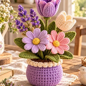 May include: A crochet flower arrangement in a purple pot featuring a purple tulip, a cream tulip, a lavender sprig, a light purple daisy, and a pink daisy. Each flower has green crocheted leaves and stems. The pot has a cream-coloured textured rim.
