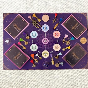 May include: A purple board game titled The Forgiveness Spell for You with four decks of cards, a central chakra diagram, and various metal charms including a dreamcatcher, skull, and cross. The board features geometric patterns and gold accents.