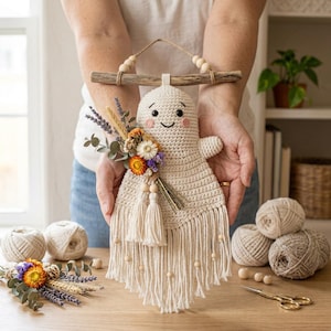 May include: A cream-colored crocheted ghost wall hanging suspended from a wooden branch with a jute cord. The ghost features embroidered black eyes, a smile, and pink cheeks. It is adorned with a dried flower bouquet and long fringe.