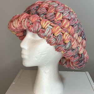 May include: A crochet bucket hat featuring a textured puff stitch pattern. The yarn is a variegated blend of coral pink, soft yellow, and grey tones, creating a multi-colored appearance throughout the entire structure of the hat.