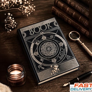 May include: A black hardback book titled THE BOOK with intricate silver geometric and circular patterns on the front cover. The design features celestial-inspired symbols, lines, and text. The book is positioned alongside a magnifying glass and a pen.