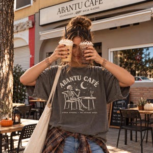 May include: A charcoal gray t-shirt featuring a white graphic of a skeleton sitting at a table with a coffee cup, a crescent moon, and stars. The text reads ABANTU CAFE and GOOD COFFEE NO EXPIRY DATE. The shirt has short sleeves and a crew neckline.