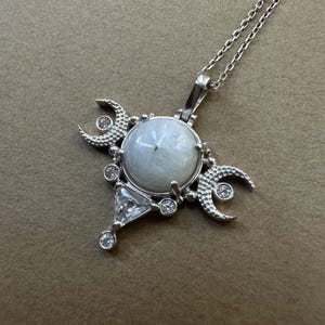 May include: A silver-toned pendant necklace featuring a central round white moonstone cabochon. The setting includes two textured crescent moon shapes, a triangular clear gemstone, and several small round clear accent stones.