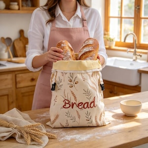 May include: A cream-colored fabric bread bag featuring a botanical print of wheat stalks and olive branches, with the word Bread embroidered in brown. The bag has a light yellow interior lining, a drawstring closure with black toggles, and contains two loaves of crusty bread.