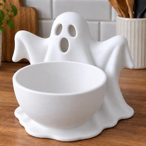 Pode incluir: Uma tigela de cerâmica branca integrada com uma figura de fantasma esculpida com dois olhos ovais e uma boca oval. O fantasma tem braços estendidos e uma base fluida, proporcionando uma base estável para a tigela circular frontal.
