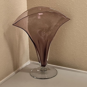 Pode incluir: Um vaso de vidro malva translúcido com um topo largo, alargado e em forma de leque, e um corpo estreito e canelado que se afunila para uma base circular transparente. O vidro apresenta um acabamento liso e polido com variações.