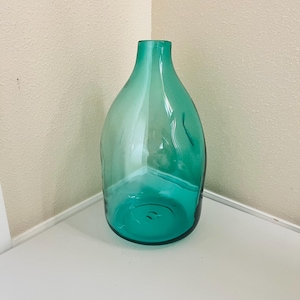 Pode incluir: Um vaso de vidro translúcido em tom verde-azulado com um corpo arredondado e bulboso e um gargalo estreito e aberto. O vidro apresenta subtis reentrâncias orgânicas na superfície, criando uma silhueta fluida e assimétrica que capta a luz.
