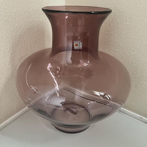 Pode incluir: Um vaso de vidro malva translúcido com um corpo largo e arredondado e um pescoço alargado. O vaso apresenta um autocolante transparente no pescoço com o texto BLENKO HANDMADE e um logótipo circular laranja. O vidro é liso e polido.